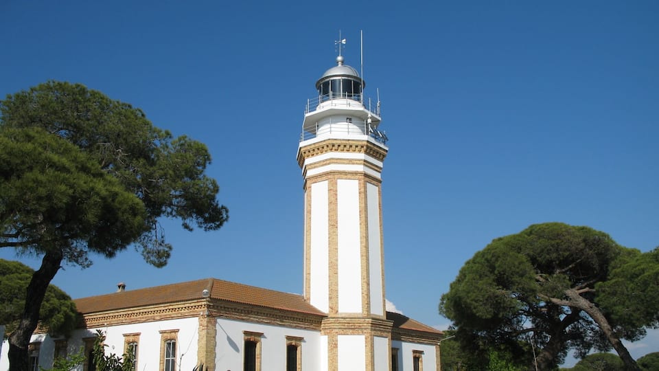 A lighthouse with a white and beige facade, a red-tiled roof, and a weather vane on top.