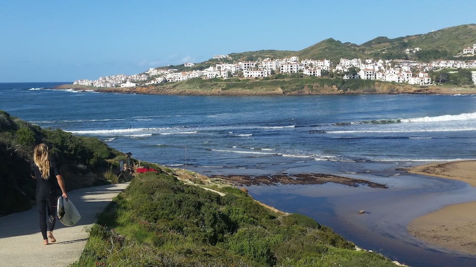 A coastal town with white buildings on a hillside, a sandy beach, and waves crashing onto the shore.