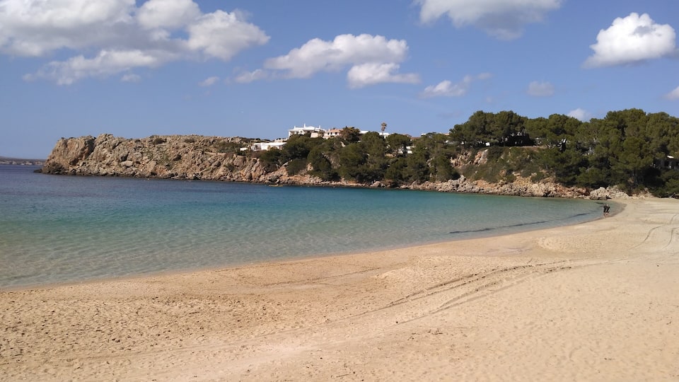 A beach with clear turquoise water, a sandy shore, and a rocky cliff with buildings on top.