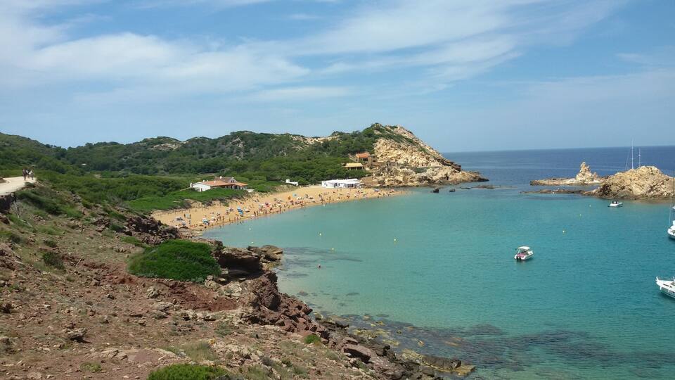 A coastal landscape with a sandy beach, rocky cliffs, and clear turquoise waters.
