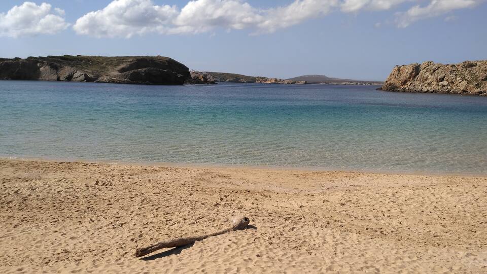 A sandy beach with clear blue water and rocky cliffs in the background.