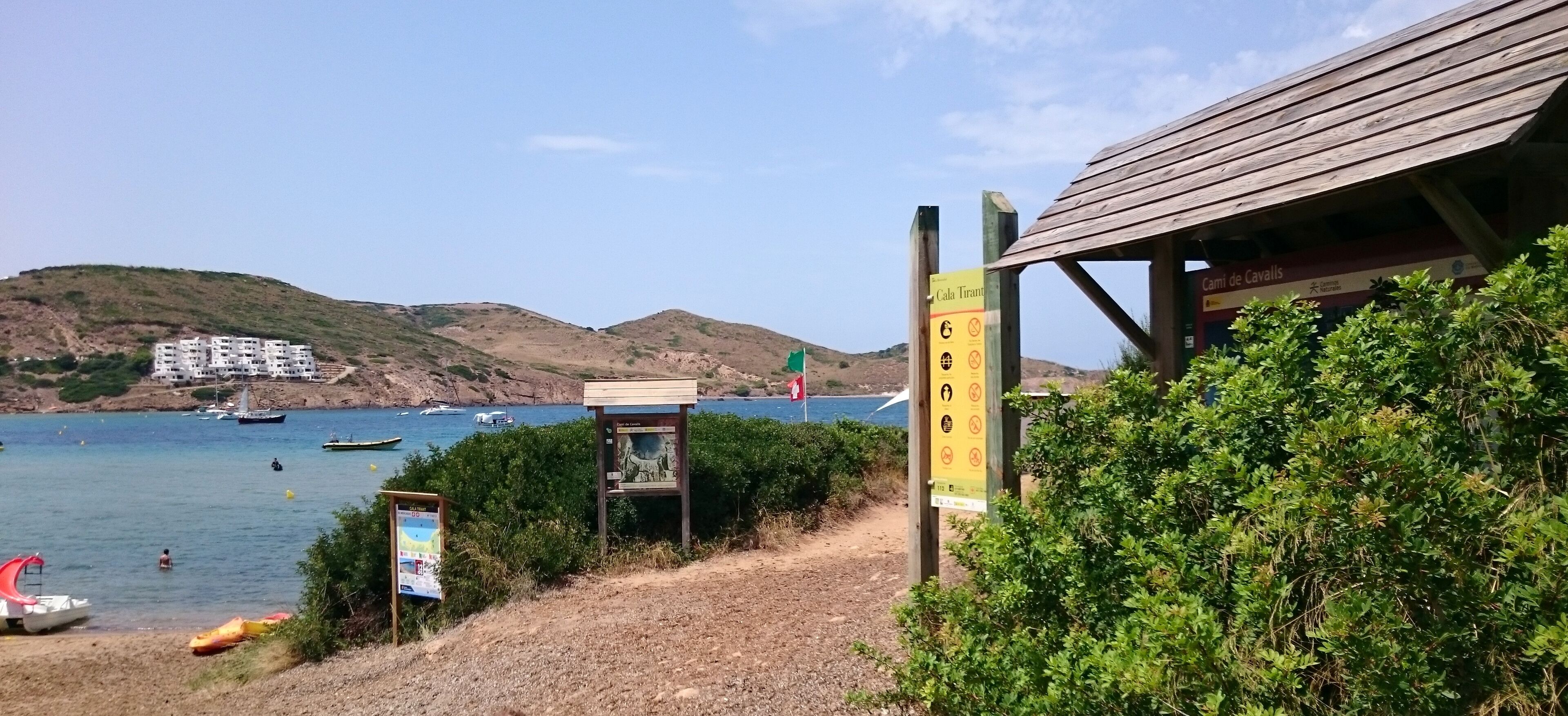 A coastal path with a wooden shelter, a signboard, and a view of boats on the water.