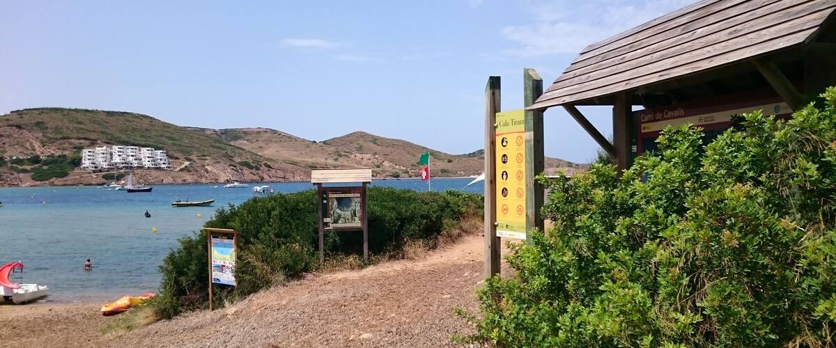 A coastal path with a wooden shelter, a signboard, and a view of boats on the water.
