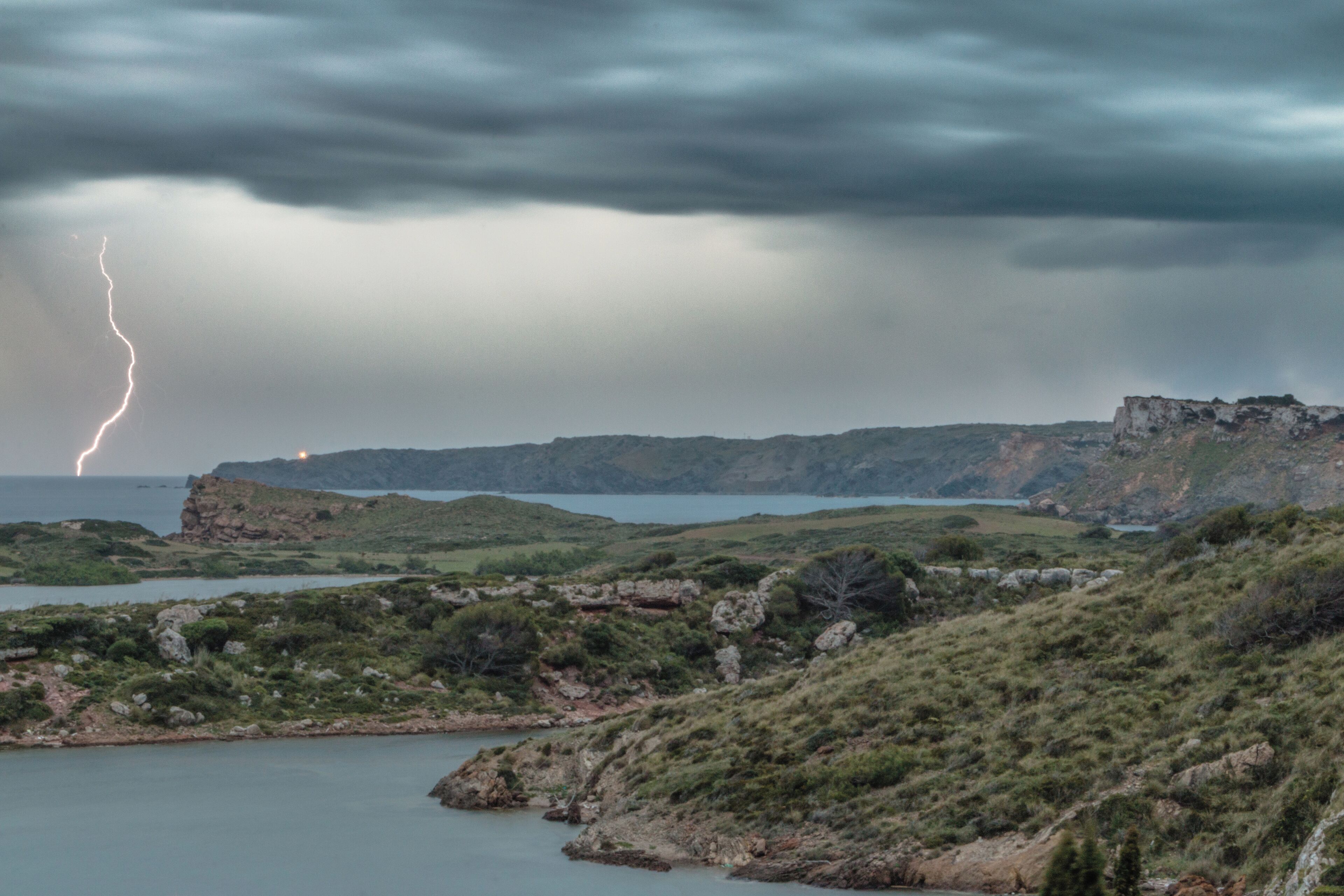 A coastal landscape with rocky terrain, a body of water, and a distant lightning strike.