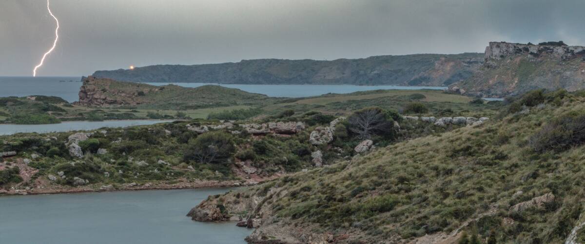 A coastal landscape with rocky terrain, a body of water, and a distant lightning strike.