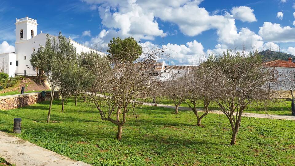 A white church with a bell tower, a stone wall, and a grassy area with trees.