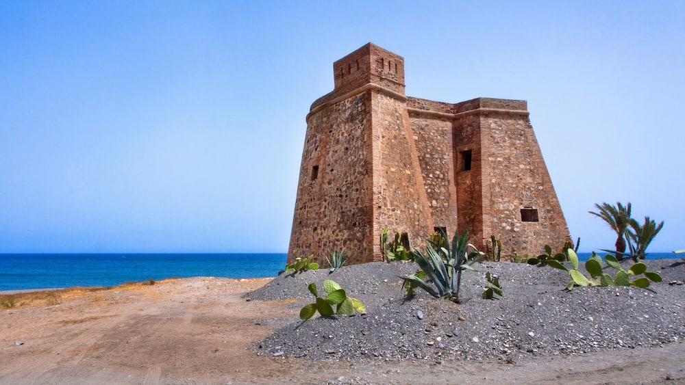 Un fort situé sur la côte, offrant une vue dégagée sur la mer, avec un paysage sablonneux en premier plan parsemé de cactus.
