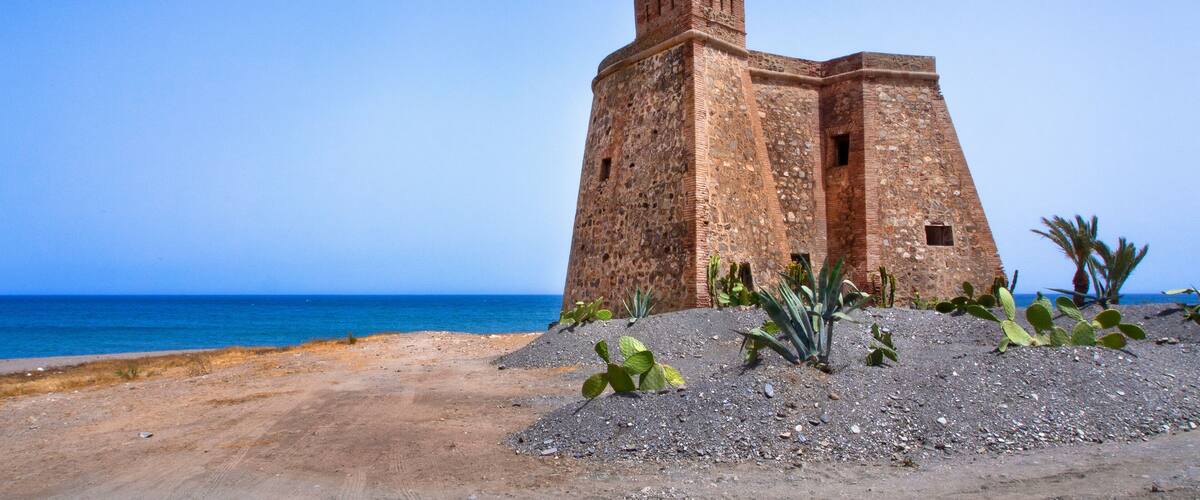 Un fort situé sur la côte, offrant une vue dégagée sur la mer, avec un paysage sablonneux en premier plan parsemé de cactus.