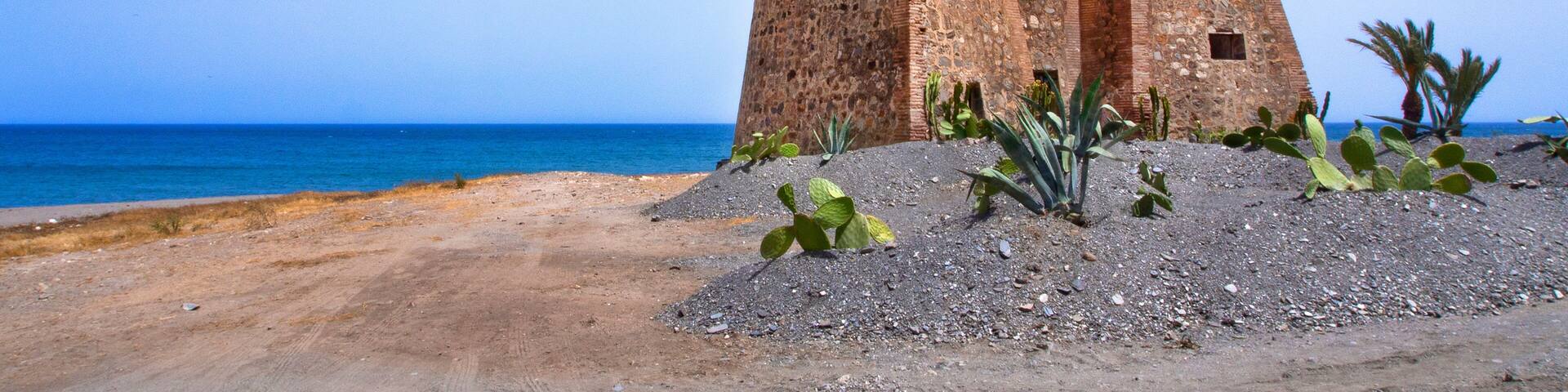 A coastal fort with a clear view of the sea and a sandy foreground with cacti.