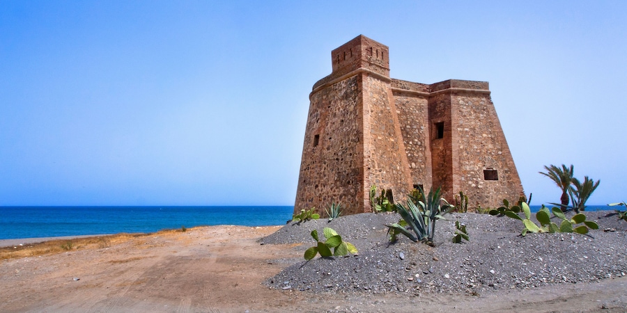 Un fort situé sur la côte, offrant une vue dégagée sur la mer, avec un paysage sablonneux en premier plan parsemé de cactus.