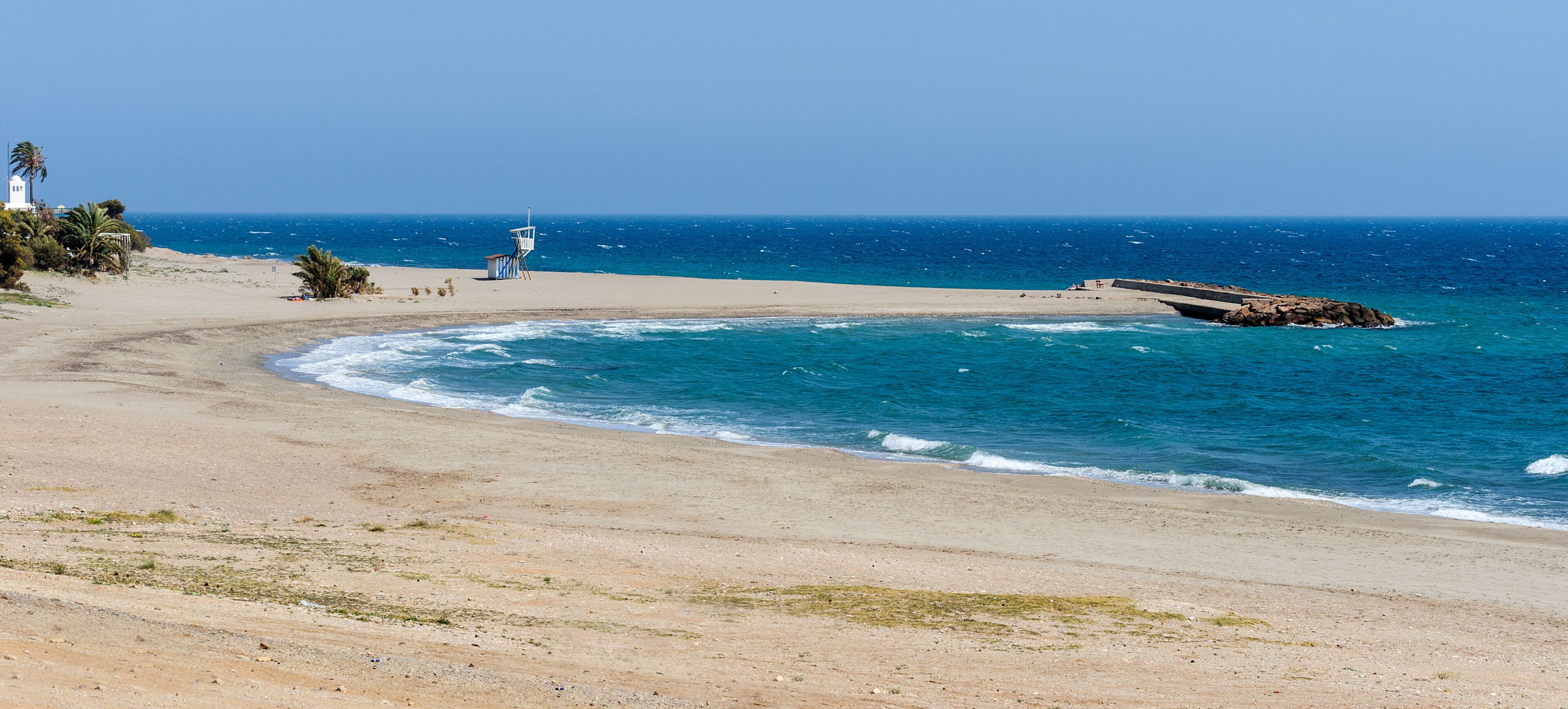 A beach with a lifeguard tower, a rocky breakwater, and a clear blue sky.