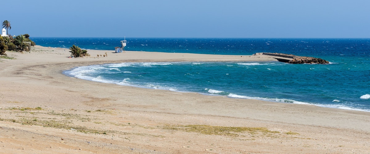 A beach with a lifeguard tower, a rocky breakwater, and a clear blue sky.