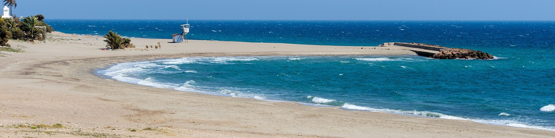 A beach with a lifeguard tower, a rocky breakwater, and a clear blue sky.