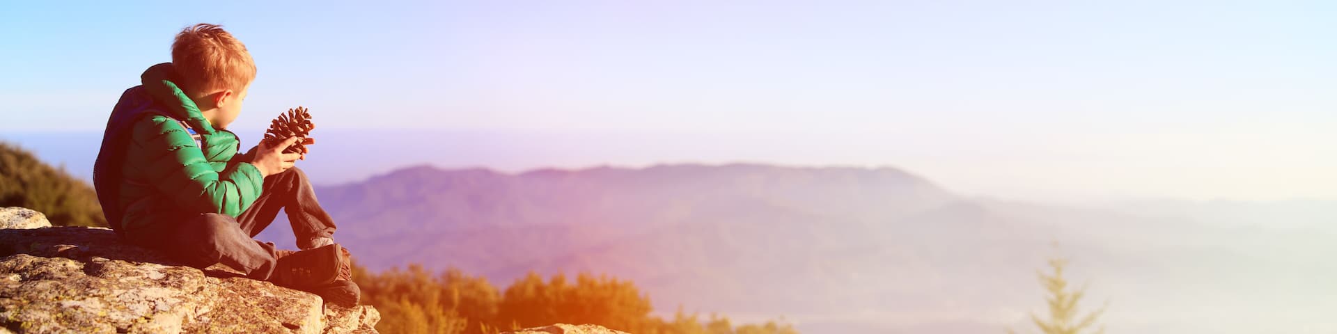 little boy hiking in scenic mountains looking at pine cone