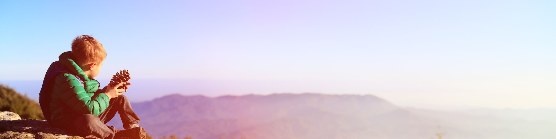 little boy hiking in scenic mountains looking at pine cone