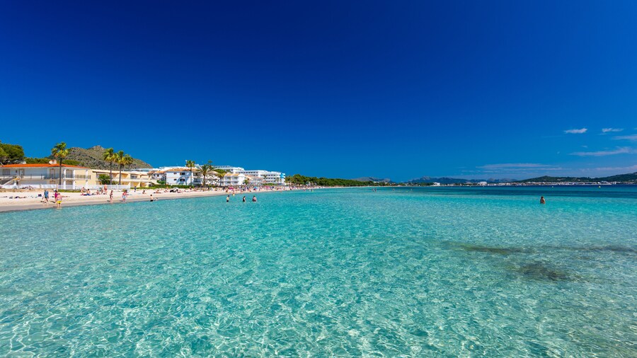 Un mar turquesa cristalino, una playa arenosa y un resort con edificios blancos y palmeras.
