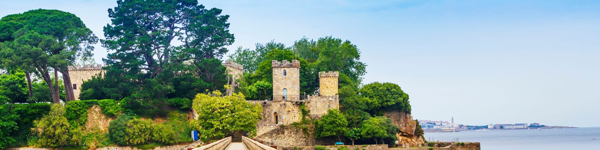 A wooden bridge leading to a historic stone castle surrounded by lush greenery and overlooking a calm body of water.