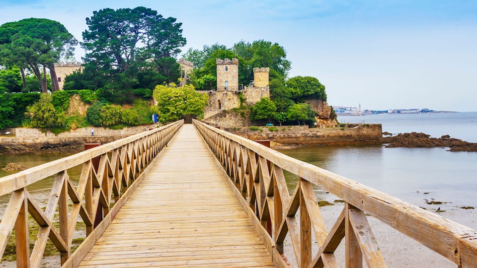 A wooden bridge leading to a historic stone castle surrounded by lush greenery and overlooking a calm body of water.