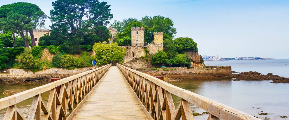 A wooden bridge leading to a historic stone castle surrounded by lush greenery and overlooking a calm body of water.