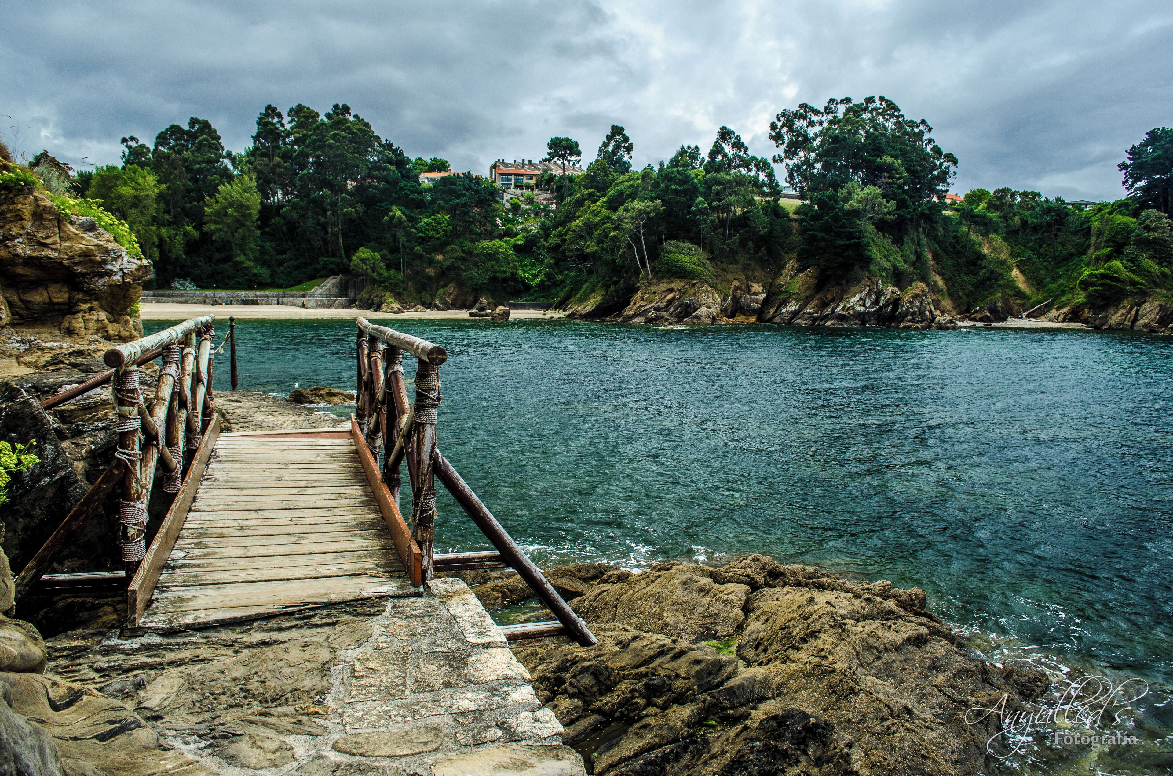 A wooden bridge leading to a beach with a rocky shoreline and a house in the background.