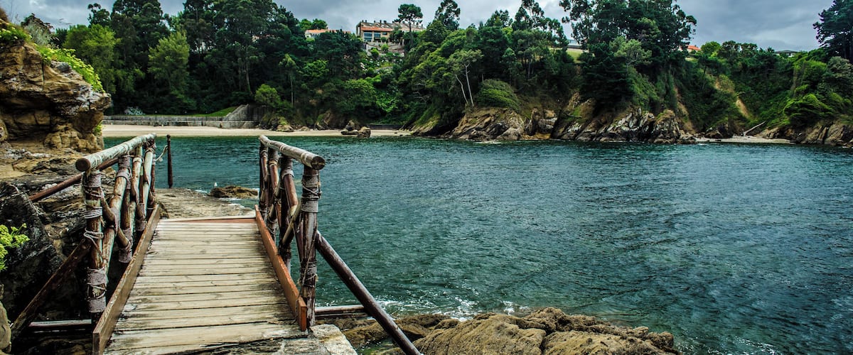 A wooden bridge leading to a beach with a rocky shoreline and a house in the background.