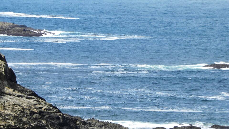 A coastal landscape with rocky shores, waves crashing against the rocks, and a lighthouse on a hill in the distance.