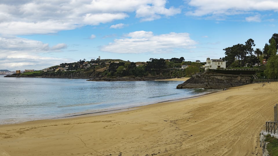 A sandy beach with a rocky outcrop and a white building on a hill.