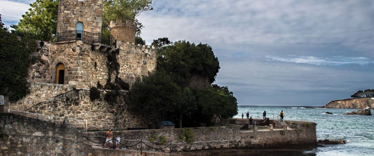 A historic stone castle with a tower, situated by the sea with a rocky coastline in the background.