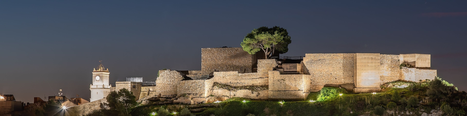 Una fortaleza de piedra histórica con una torre del reloj, iluminada por luces artificiales, rodeada de abundante vegetación.