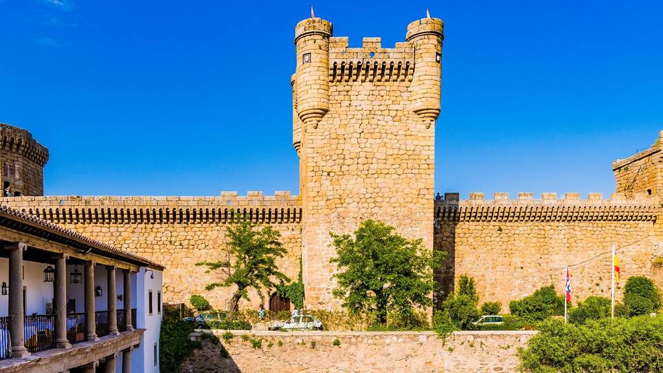 A historic stone castle with two prominent towers, a white building with columns in the foreground, and lush greenery.