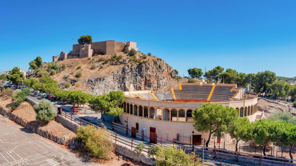 A coastal town with a historic fort on a hill, a round building with arches, and a clear blue sky.