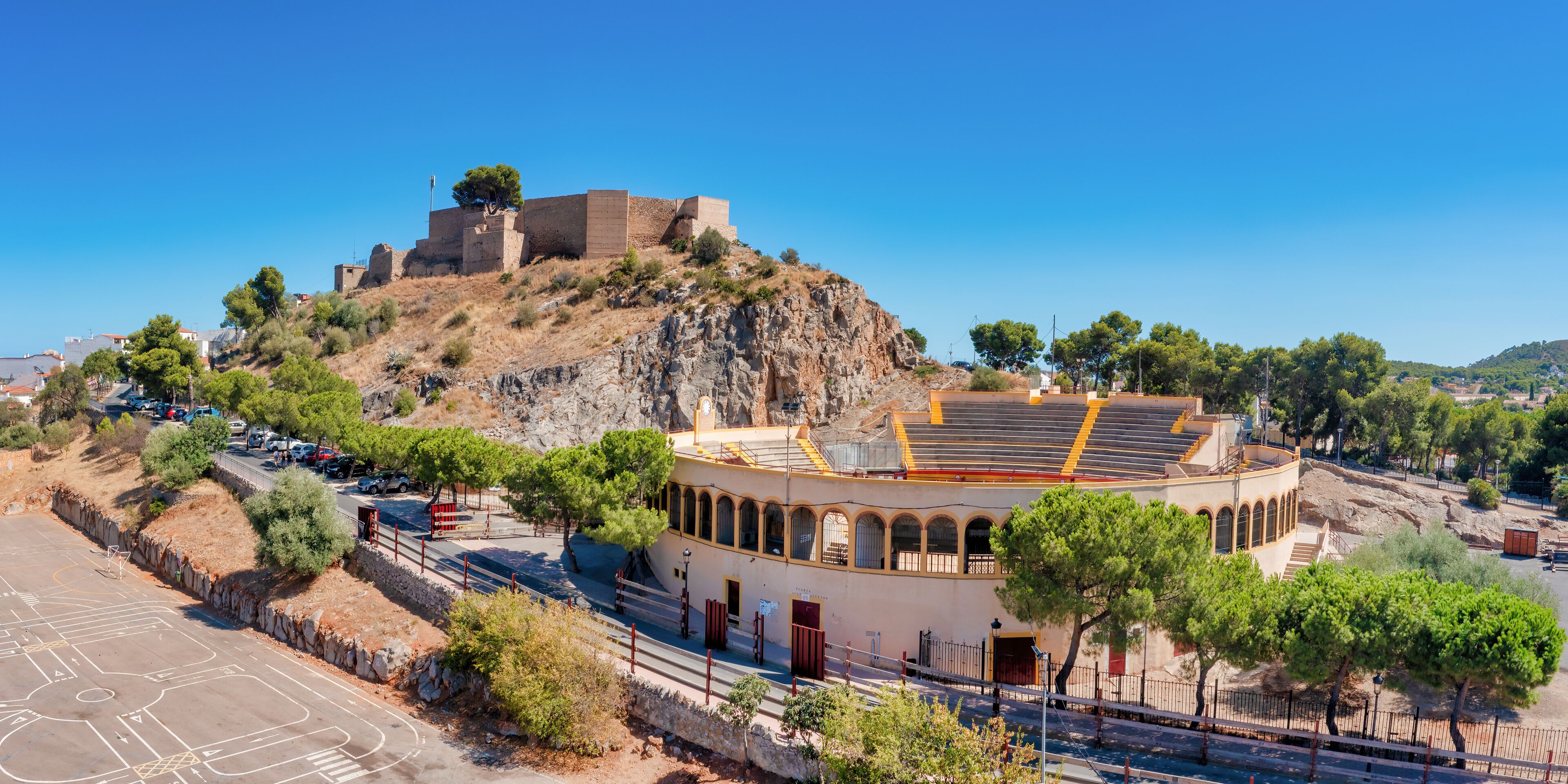 A coastal town with a historic fort on a hill, a round building with arches, and a clear blue sky.