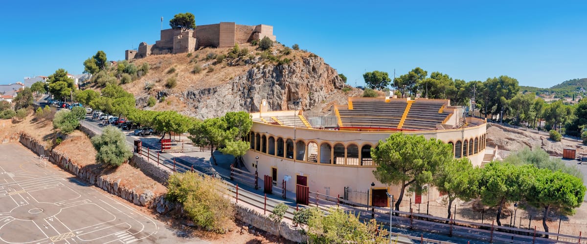 A coastal town with a historic fort on a hill, a round building with arches, and a clear blue sky.
