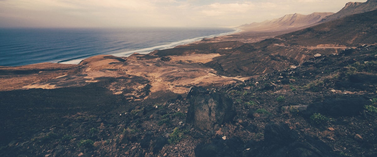 A coastal landscape with a rocky shoreline, a distant view of the ocean, and a clear sky with scattered clouds.
