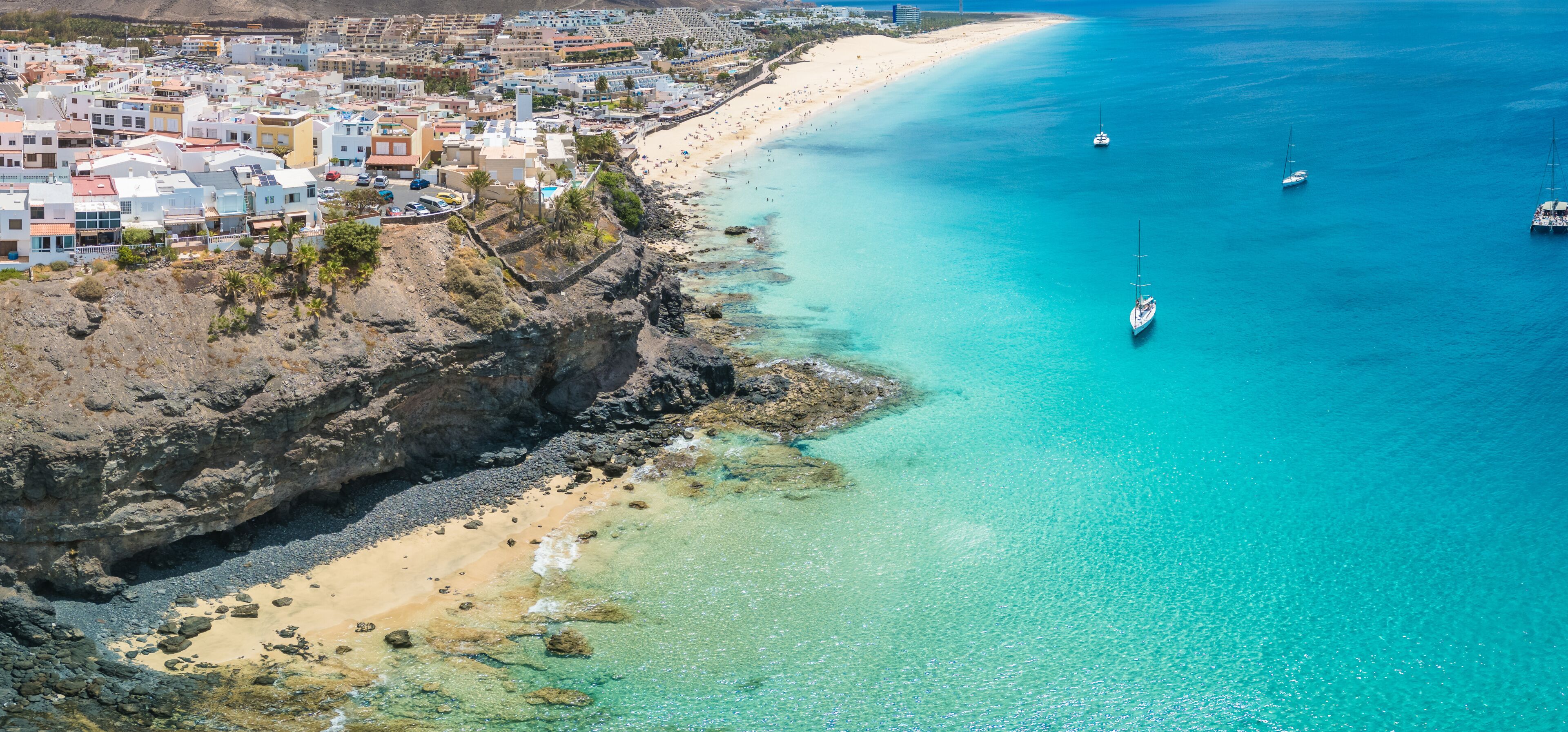 Un pueblo costero con una playa arenosa, aguas turquesas cristalinas y varios veleros.