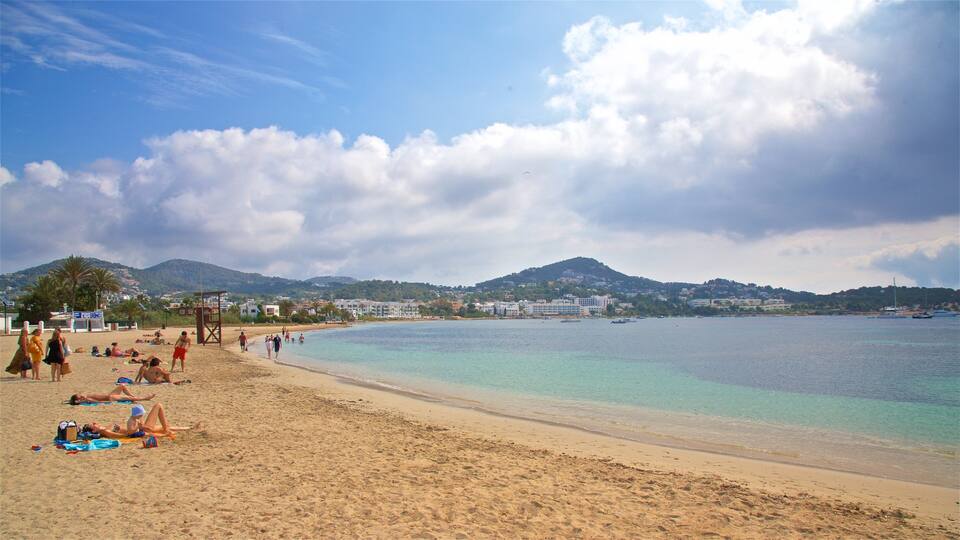 A beach with clear turquoise waters, sandy shore, and a backdrop of hills and buildings.