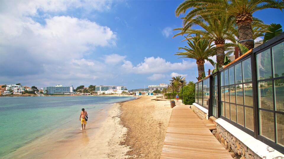 A beach with clear turquoise water, a wooden walkway, palm trees, and buildings in the background.