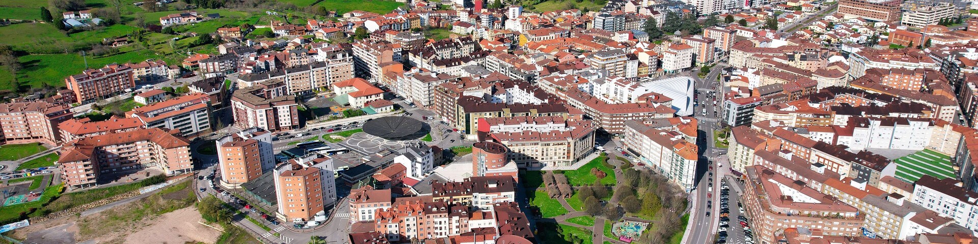 Vista aérea de una ciudad con un edificio circular destacado, rodeado de construcciones residenciales y comerciales, y colinas verdes al fondo.