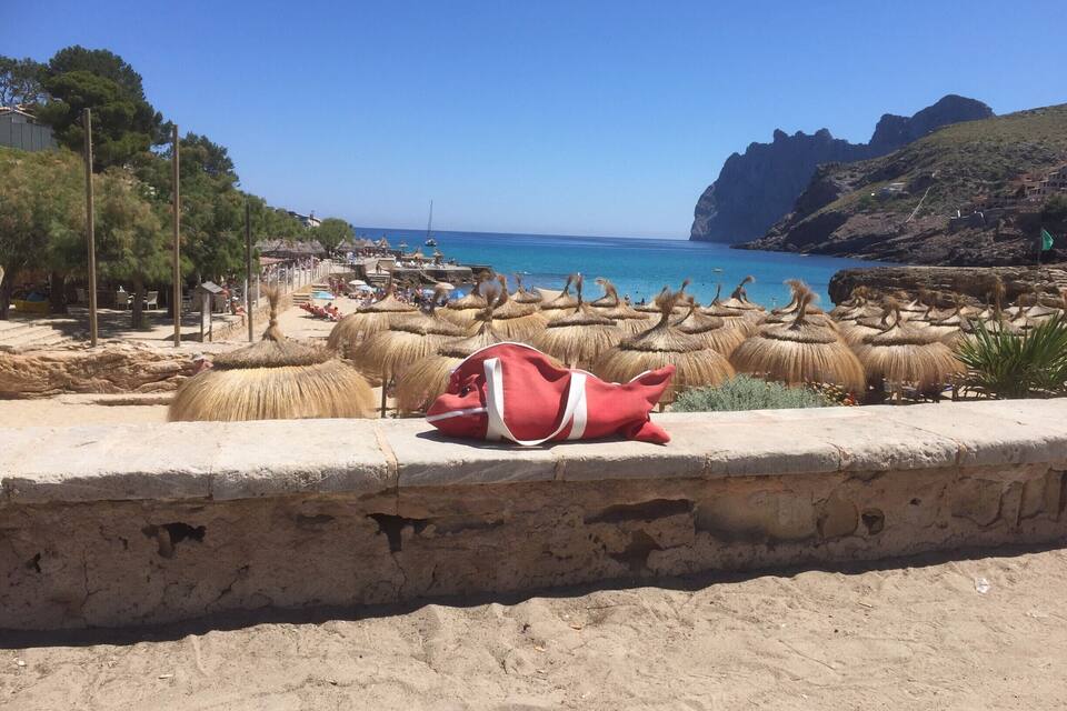 Une plage avec des parasols en chaume, un sac rouge et une falaise rocheuse en arrière-plan.