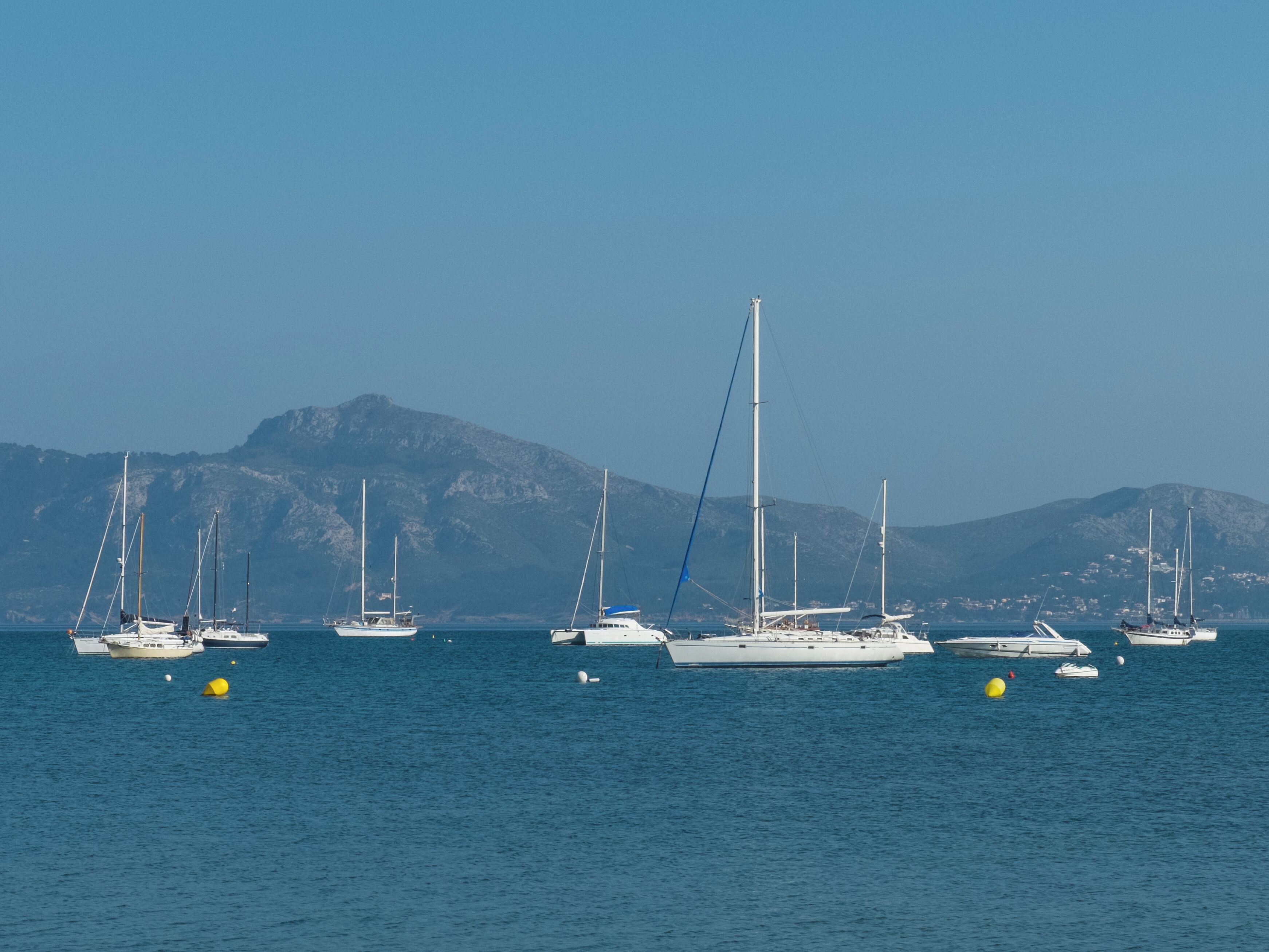 A marina with several sailboats anchored in calm waters, with a mountainous backdrop.