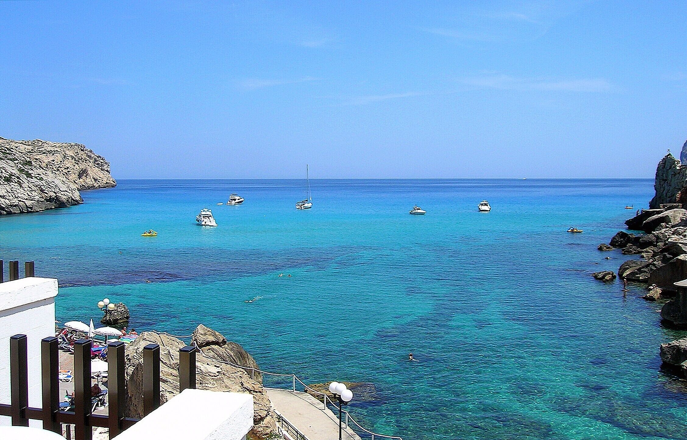 A coastal scene with clear turquoise waters, rocky cliffs, and several boats anchored near the shore.