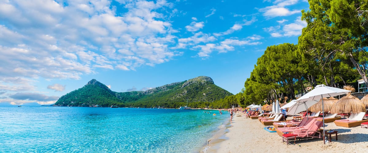 A beach with clear turquoise water, a sandy shore, and a mountain in the background.
