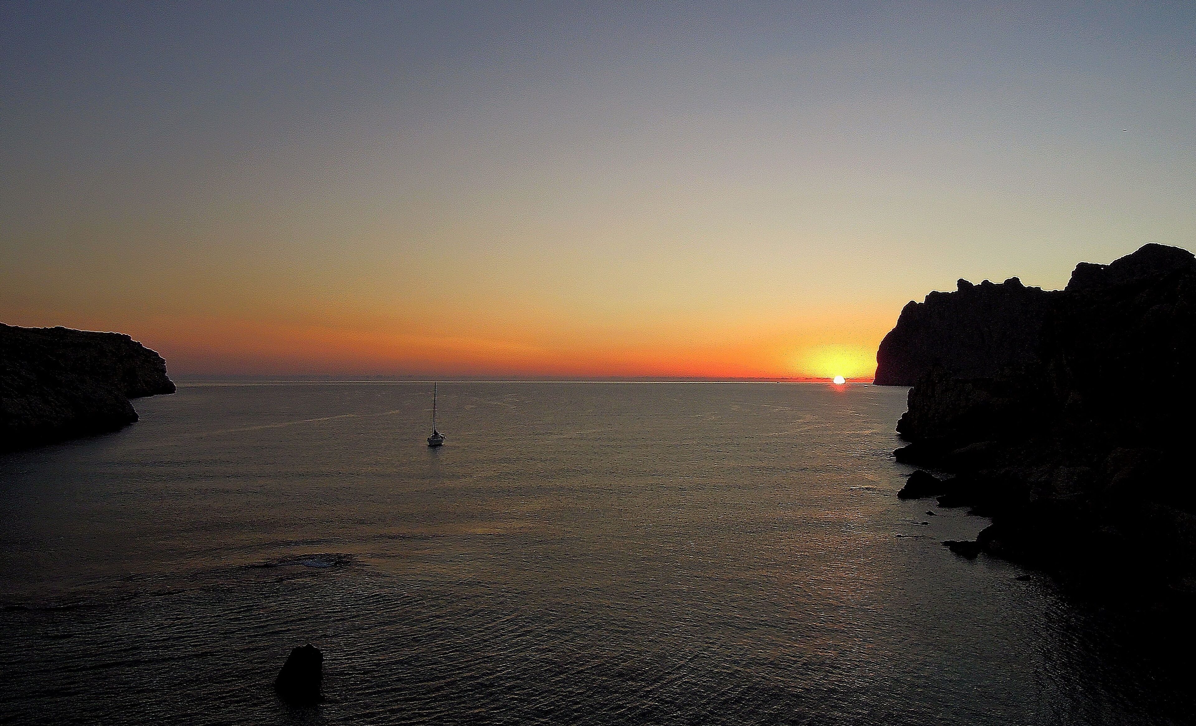 A sunset over a calm sea with a sailboat and rocky cliffs.