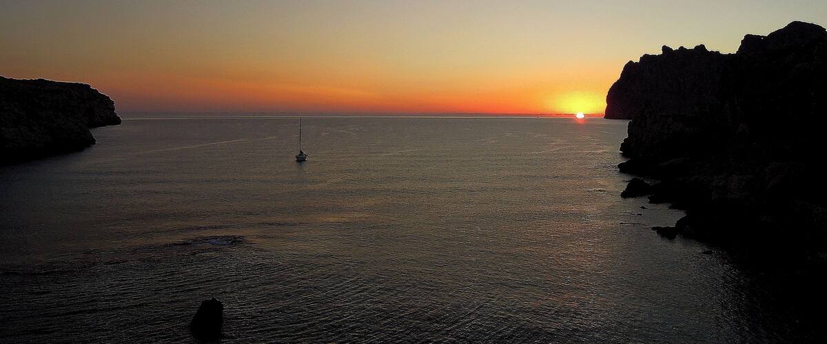A sunset over a calm sea with a sailboat and rocky cliffs.