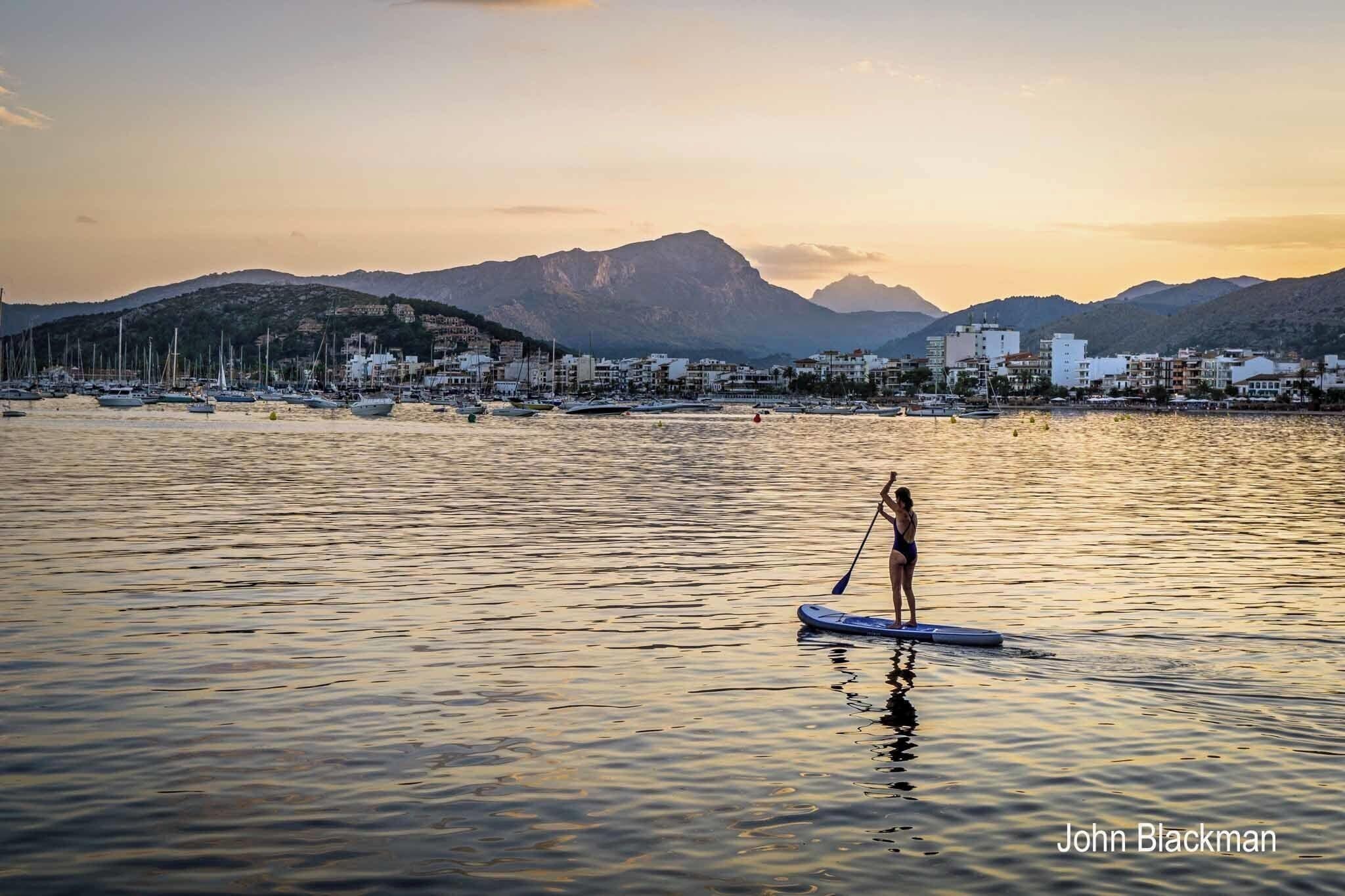 A person paddleboarding on calm waters with a backdrop of mountains and a coastal town.