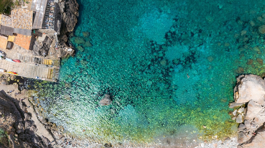 Aerial view of Cala Deia with clear turquoise water, rugged rocky cliffs, and people relaxing on the pebble beach Spain.