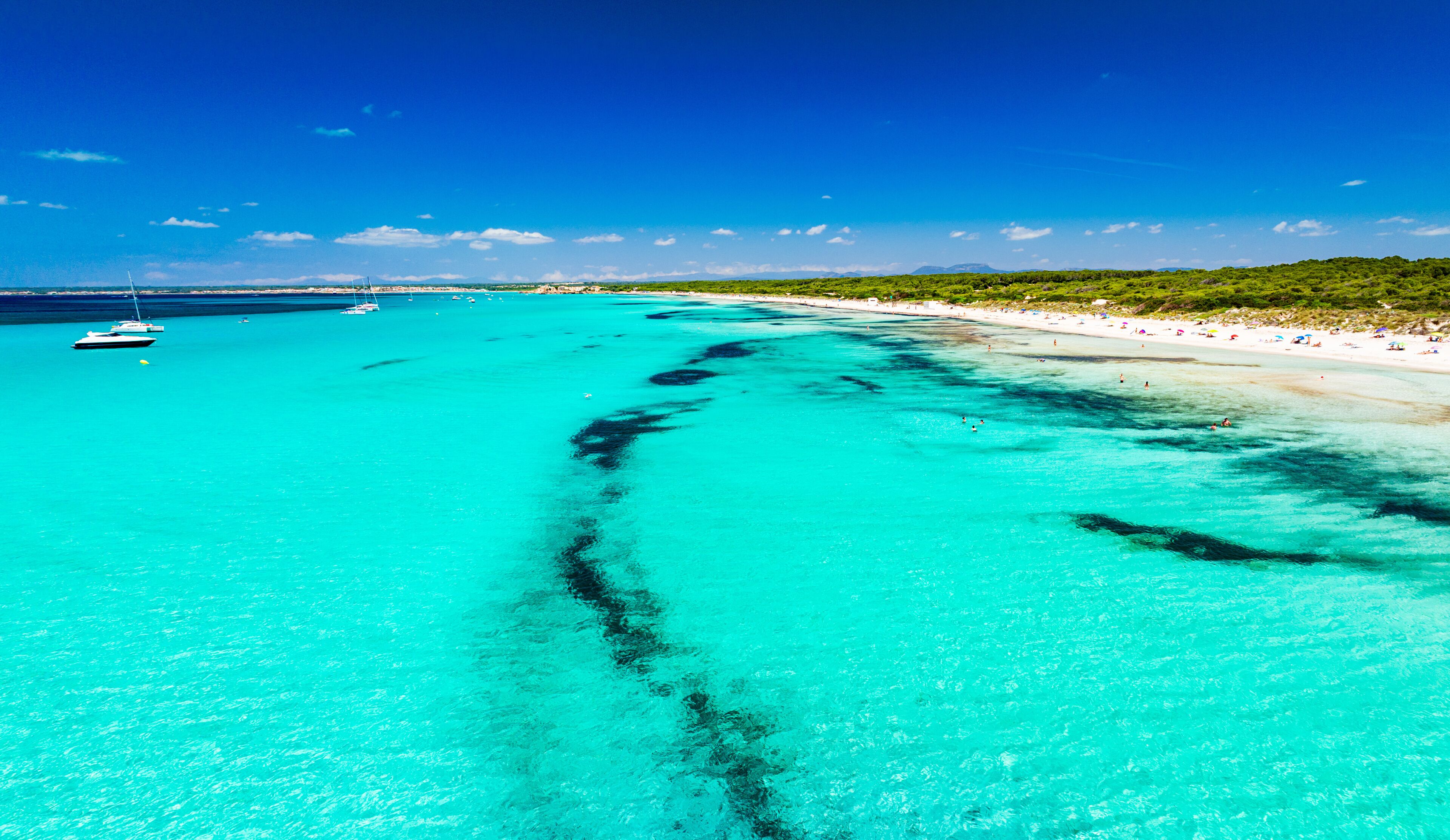 A turquoise sea with a clear underwater trail, a sandy beach, and boats in the distance.