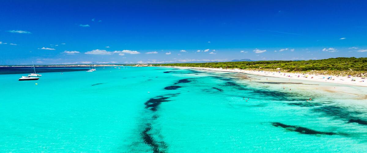 A turquoise sea with a clear underwater trail, a sandy beach, and boats in the distance.