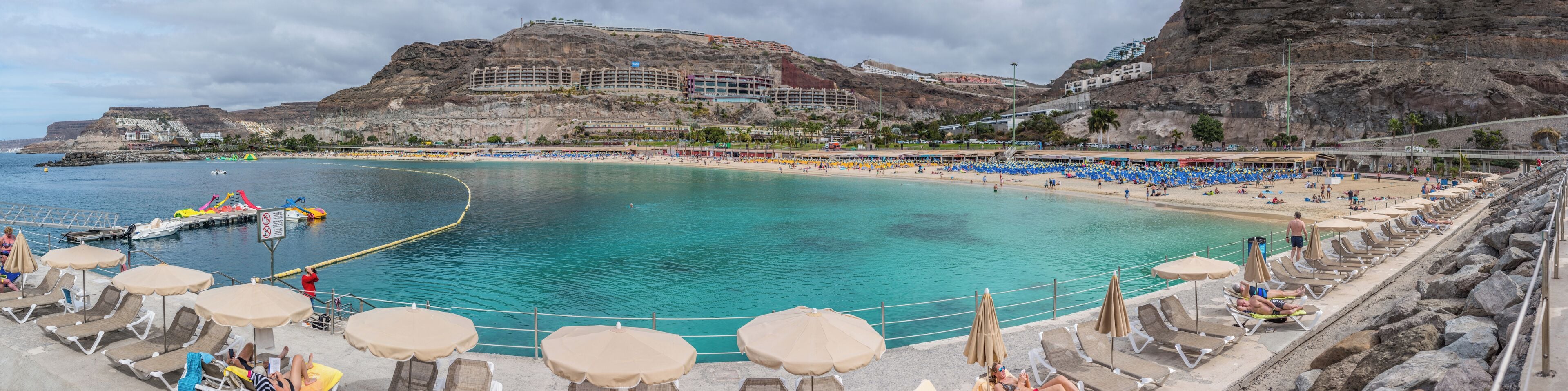 A coastal resort with a clear turquoise sea, a sandy beach, and a rocky cliff.