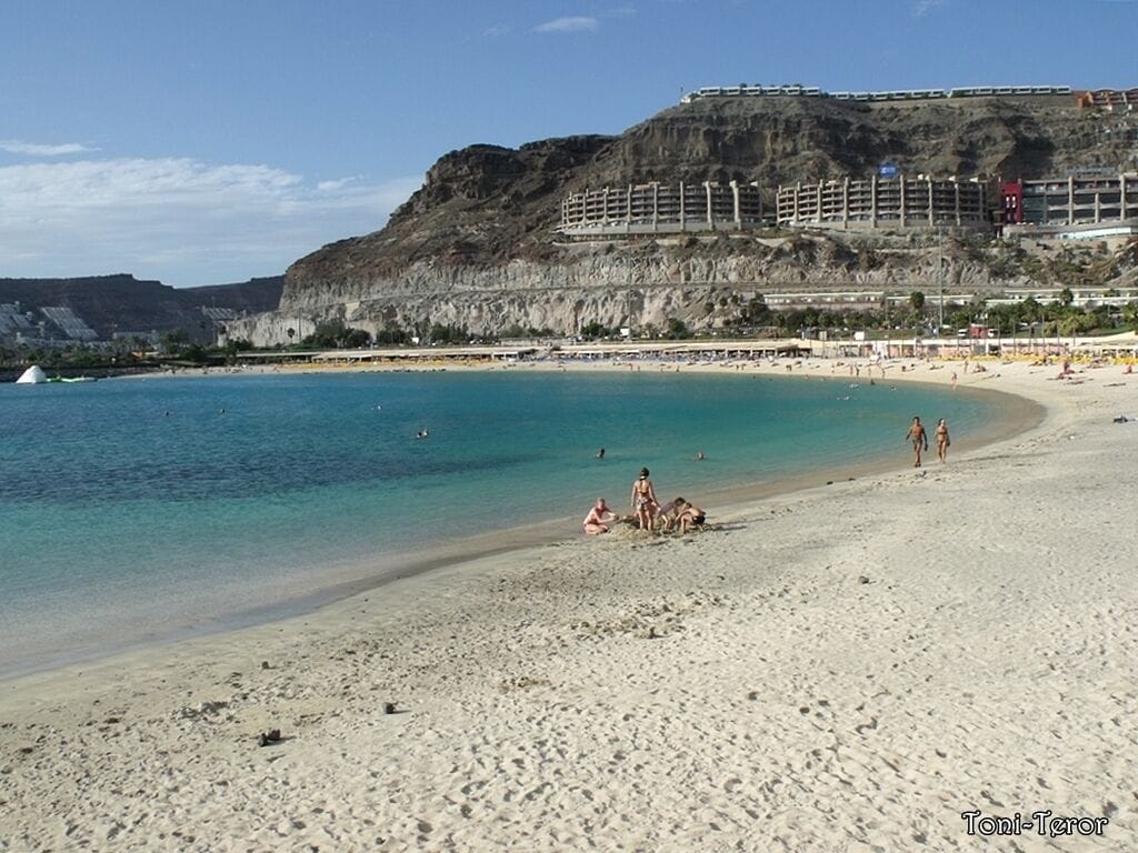 A beach with clear turquoise water, a sandy shore, and a large building on a hill in the background.